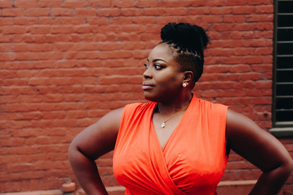 Portrait of a confident woman in a red dress posing against a brick wall.